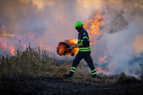Firefighter in protective gear carrying a burning log during wildfire containment efforts, with dense smoke in the background.