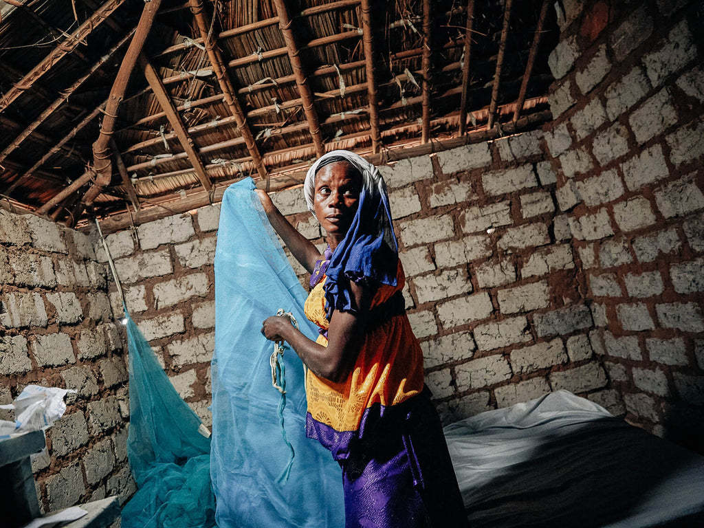 Jacqueline is holding a blue mosquito net above her bed in a small mud brick hut with a thatched roof
