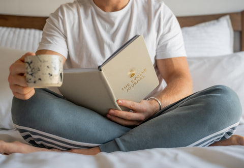 Freddie Titcombe, co-founder of Evolve Journey holding the cream Evolve Journal sat on bed holding a coffee