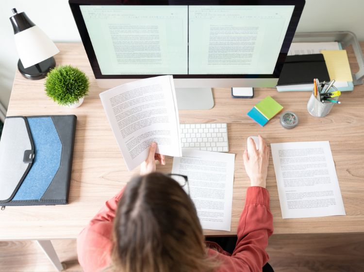 Woman at desk proofreading with screen and hard copies