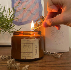 A hand uses a match to light a Boucle candle in a glass jar