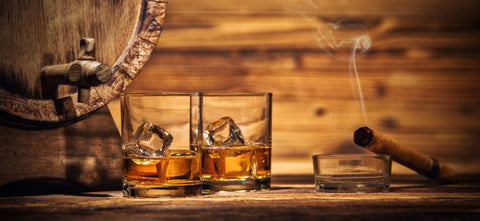 Two iced glasses of whiskey stand in front of a whiskey barrel on a wooden table. A lit cigar rests in a nearby ashtray.