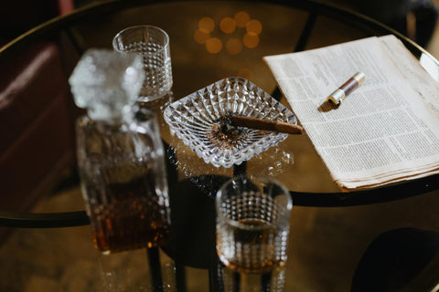 Two glasses on a glass table: one filled with whiskey, the other empty. A decanter sits between them. A lit cigar rests in an ashtray nearby. A lighter sits on a newspaper.