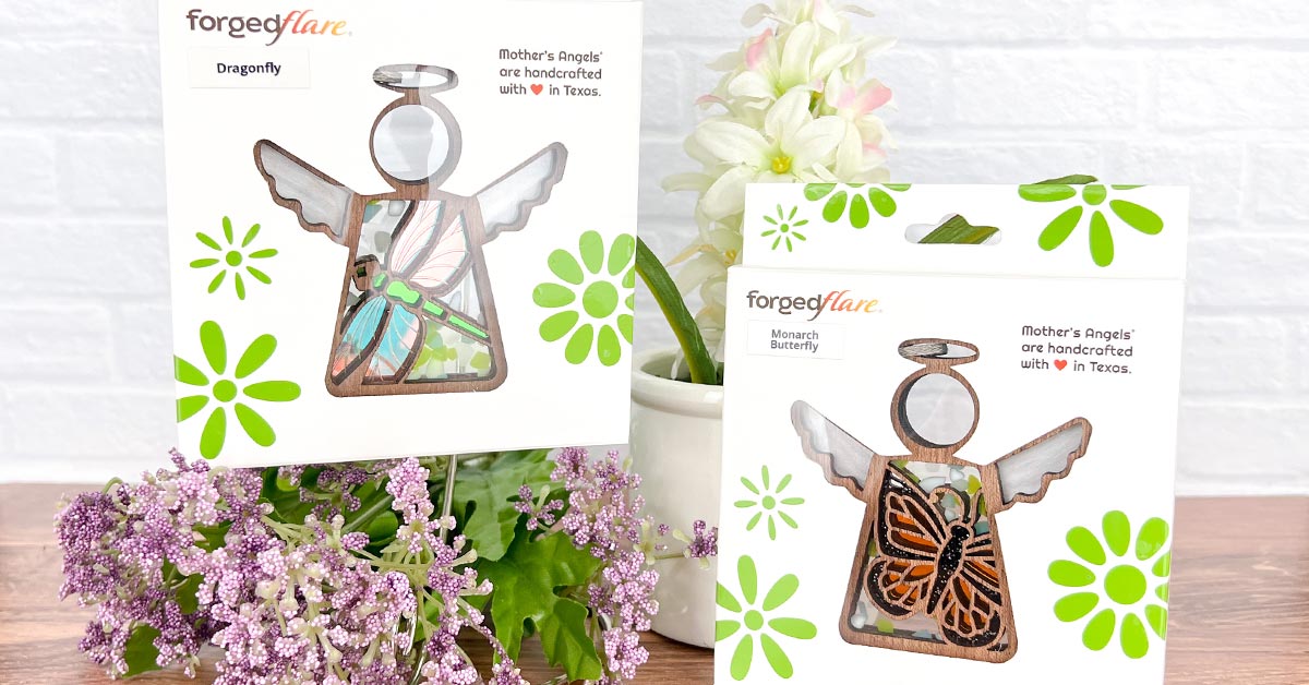 Two Forged Flare® Mother’s Angels® ornaments with dragonfly and monarch butterfly motifs, presented in packaging that promotes reusability and recycling information, highlighting the ability to recycle the packaging and its reusability. Handcrafted with love in Texas, these stained glass-inspired ornaments are displayed alongside fresh flowers, emphasizing their vibrant and natural aesthetic.