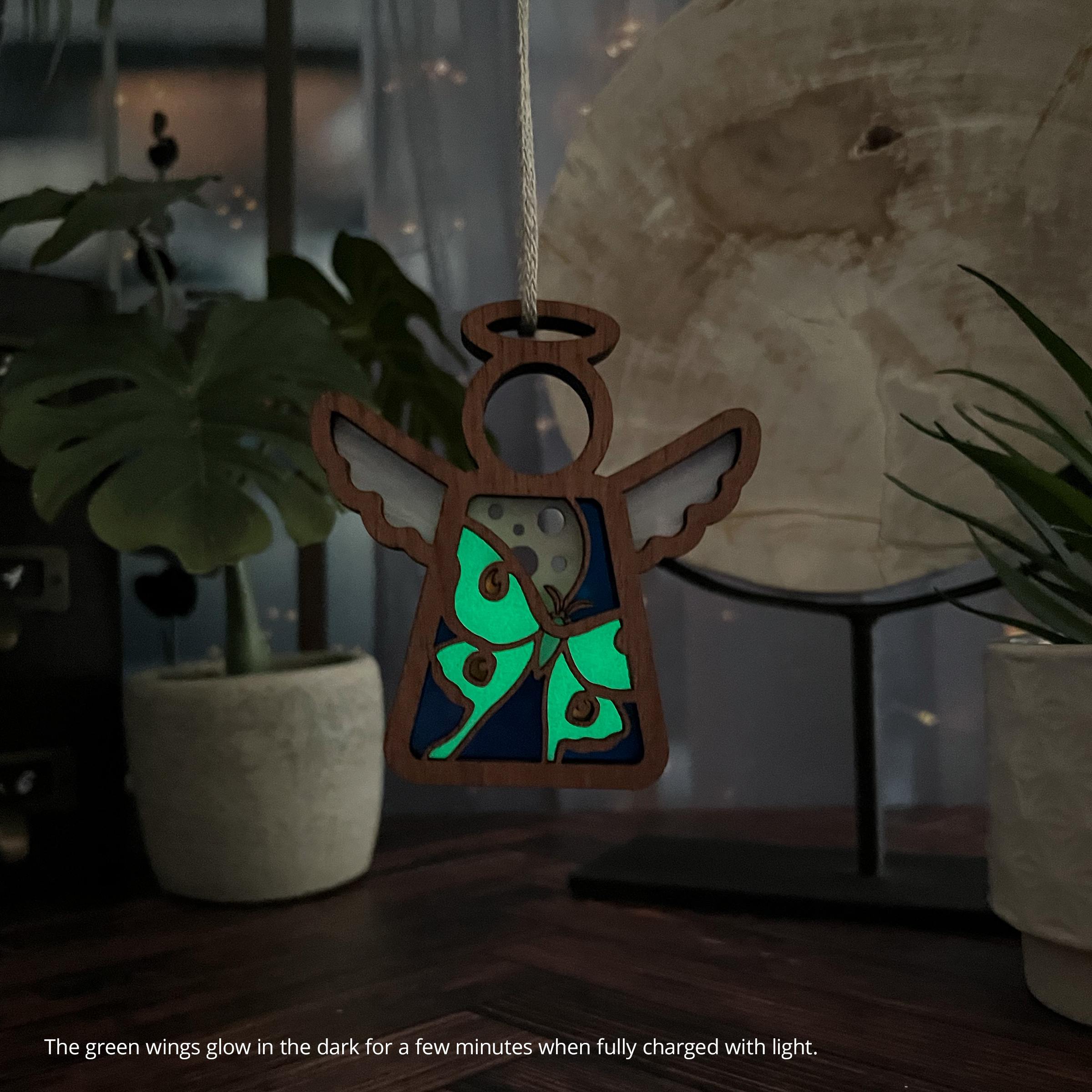 A glow-in-the-dark Luna Moth Mother’s Angels® ornament sitting on a desk in the twilight. The green wings glow in the dark for a few minutes when fully charged with light.