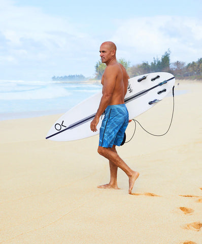 Kelly Slater walking on the beach with blue Outerknown swim trunks on and a surfboard under his right arm