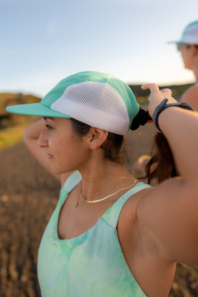 side profile of female runner with dark straight hair adjusting her ponytail while wearing her Arna Ultralight Running Cap in Celeste 