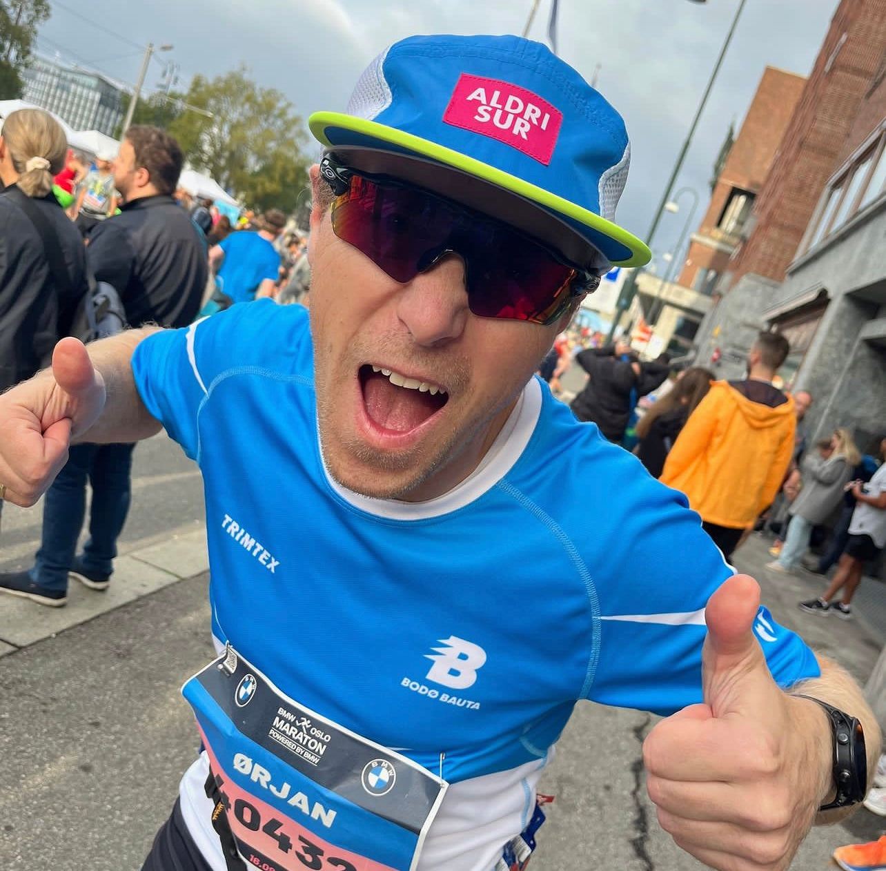 an excited Norwegian marathon road runner smiling and doing a thumbs up pose to the camera at the 2023 Oslo Marathon finish line while wearing his race bib number, a blue technical t shirt, sports sunglasses and the ALDRI SUR Arna Ultralight Running Cap in Blue
