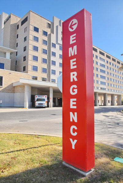 A red sign with white lettering that reads Emergency set against an exterior view of a hospital in the background.