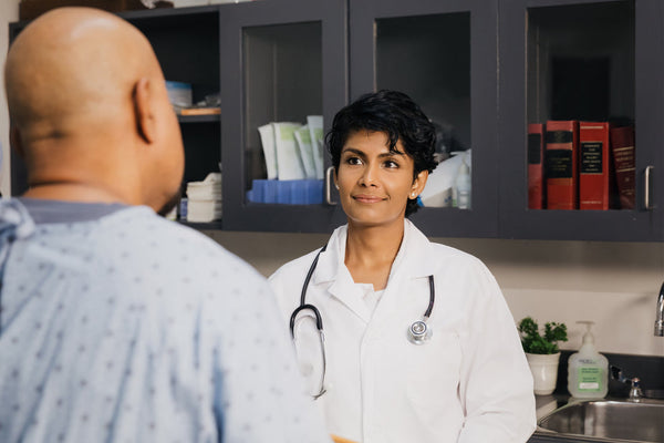 A lifestyle medicine doctor and her patient sit in the doctor's office having a conversation. She is wearing a lab coat and stethoscope, facing the camera. He is wearing a hospital gown and is facing the doctor.