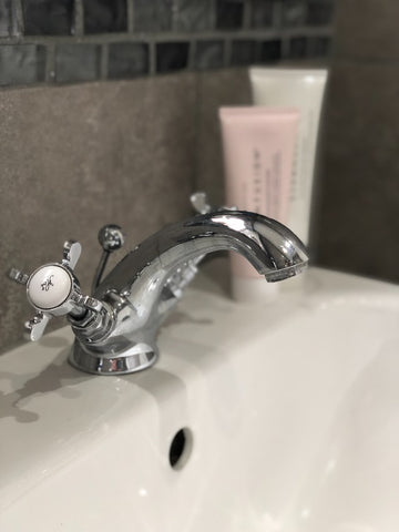 How to get rid of acne: face washing - Two tubes of gentle face cleanser sitting on a porcelain sink with an older style chrome faucet with two knobs and one spigot