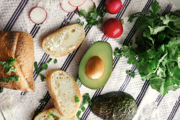 Letter vitamins: A whole avocado, a halved avocado with seed exposed plus some bread and leafy greens on a rustic tabletop. 