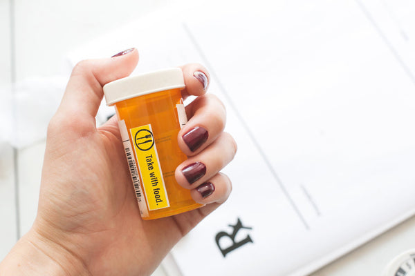 A hand holding a prescription pill bottle over a paper that says "Rx," short for prescription. Bottle has a label reading "take with food." 