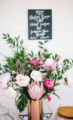 pink and hot pink flower arrangement in a gold vase with a black sign above the arrangement