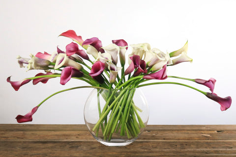 purple and white calla lilies in a large round clear vase on a wooden table
