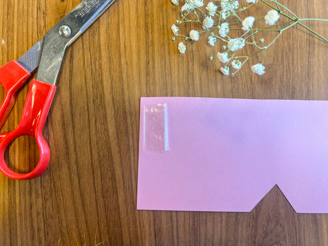 pink paper cut into a rectangle with a piece of tape on the top left corner and scissors