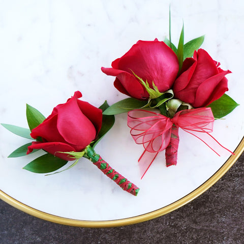 red boutonniere and corsage with red ribbon and greenery on a white table