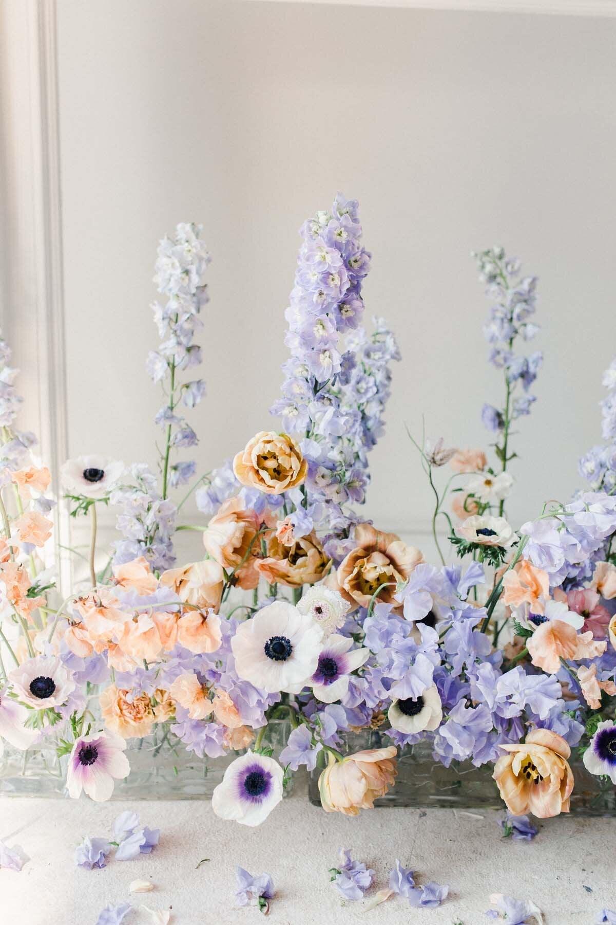 A pastel flower installment with tall periwinkle delphinium, white anemones, and peach roses
