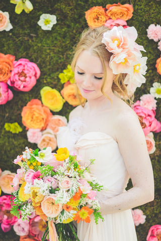 Bride in front of peony wall holding her bridal bouquet and looking down at it