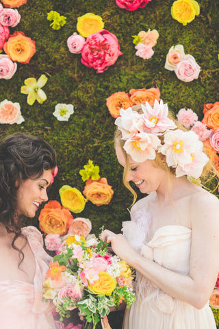 Bride and bridesmaid standing in front of peony wall looking at bridal bouquet