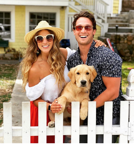 Smiling couple hugging a dog in front of a white picket fence