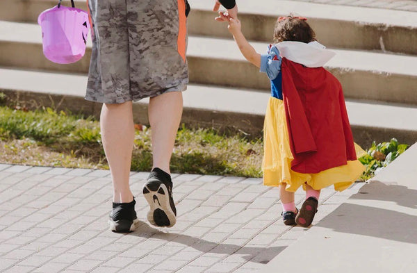 little girl dressed in a Snow White costume holding her dads hand as he holds her purple pumpkin candy basket 