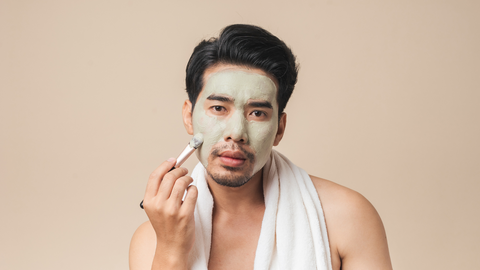 Man applying green face mask with towel around his neck. 