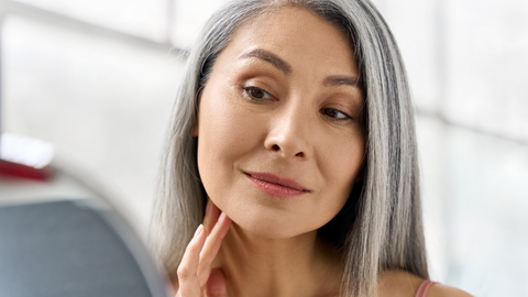 Grey haired woman looking at her skin in mirror. 