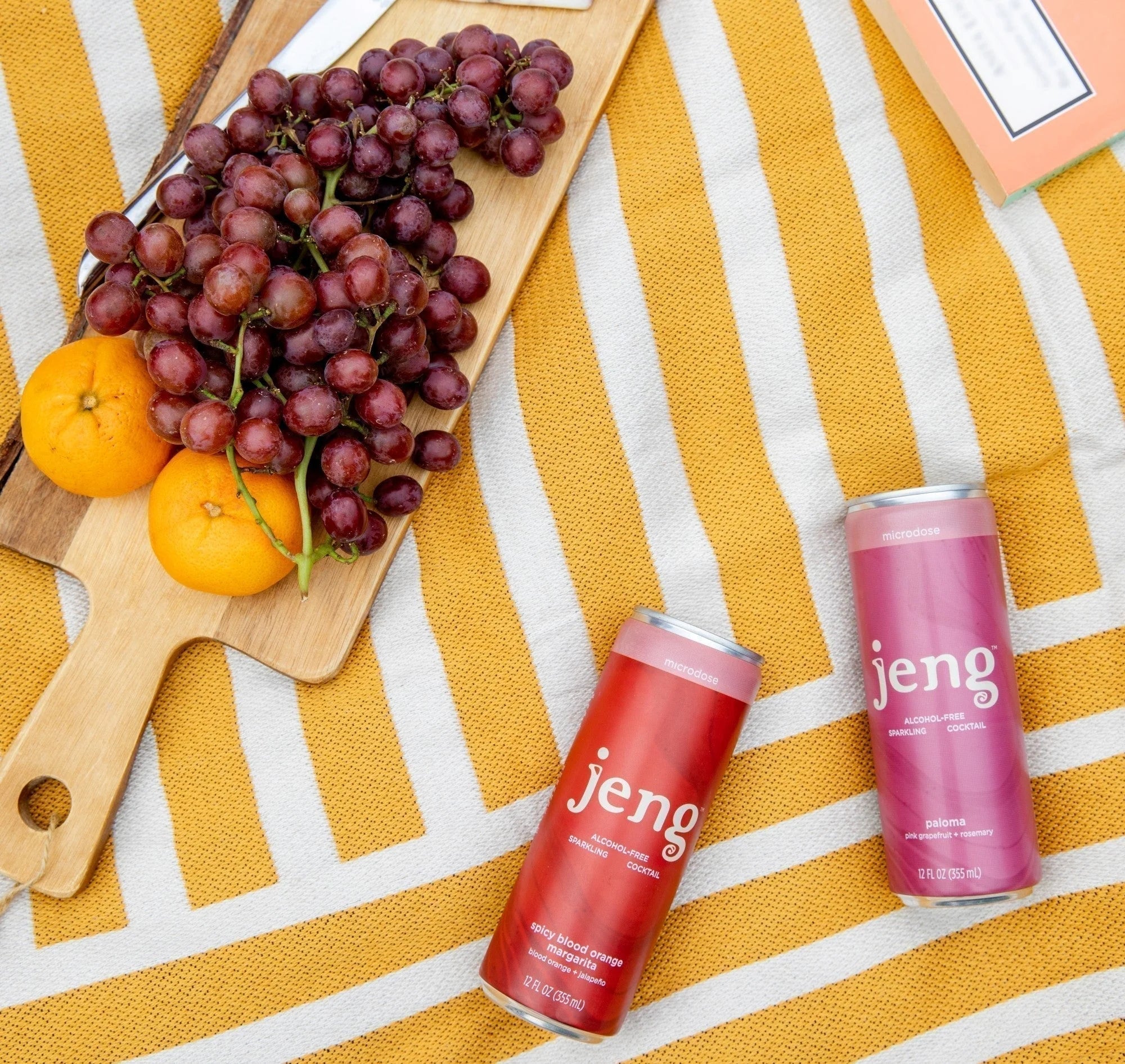 Two Jeng alcohol-free cocktails on a striped picnic blanket beside grapes and oranges, creating a vibrant, outdoor sipping setup.