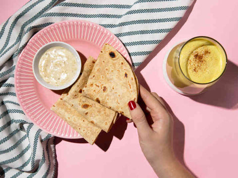 hand holding 1 roti out of 3 placed on a pink plate along with a glass of mango milkshake placed next to the plate