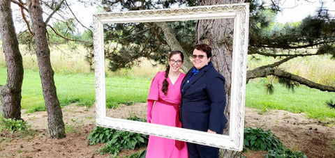 Rebecca and Erin stand in front of a tree and inside a white picture frame. Erin wears a pink dress and Rebecca wears a blue shirt with a bow tie. They are smiling.