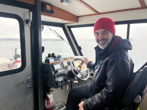 Stephen Neff wears a red toque and black rain jacket, sitting at the helm of a dive boat, holding onto the ship's wheel, smiling at the camera