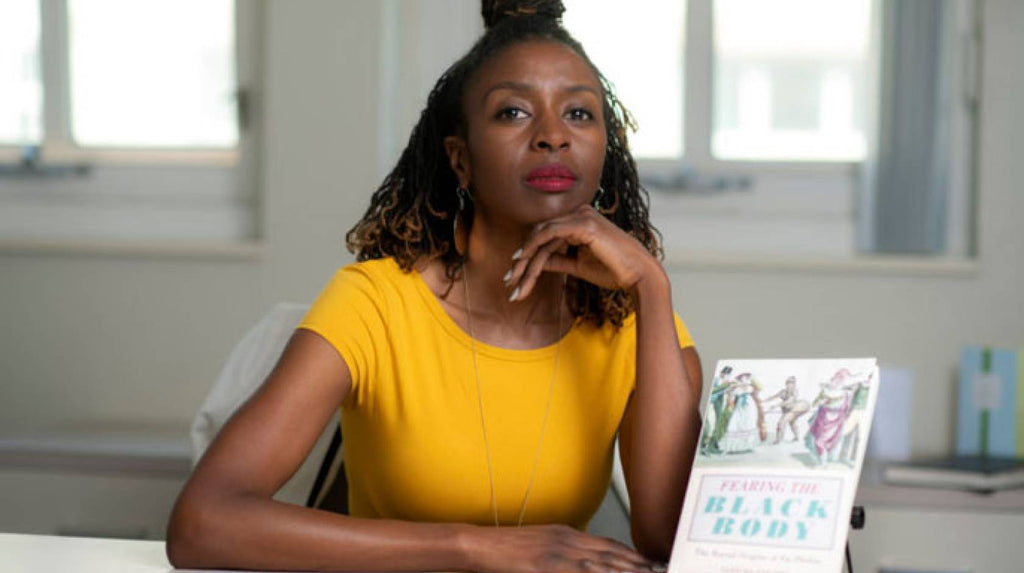 An image of UC sociology professor Sabrina Strings, Ph.D, a darkskin Black person in a yellow t-shirt, red lipstick and a necklace. In a white room, she is posed in front of her book, Fearing the Black Body: The Racial Origins of Fat Phobia. From the University of California.