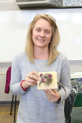 a blond light skinned wearing a grey long sleeve shirt holds her felting project. It is a felted mouse a fake taxidermy grey mouse on a wood plaque. The mouse looks like its jumping through one side of the plaque to other.