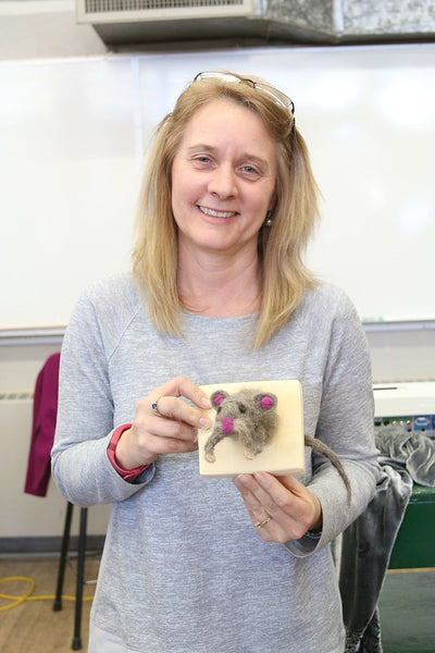 A teacher holds her felting project: a mouse on a plaque that looks like its jumping out at you! She is light skinned, blond hair, slim and smiling wearing a long sleeved grey shirt