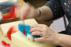 a close up of a tecahers hands working on a felting project. One hand is holding the wool and the other is using a felting needle to poke the wool into a 3D shape