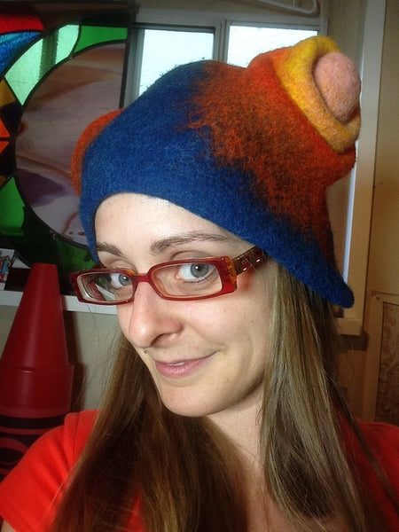 Wet Felted Hat: Zee a light skinned woman with red glasses wearing a blue red and yellow silly wet felted hat with knobby shapes on the sides