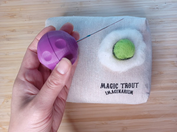 An overhead shot closeup of a hand holding a purple 3D printed felting needle holder. In the background on the table is a Magic Trout Imaginarium jute felting bag and a felting sunnyside up egg with a green yolk.