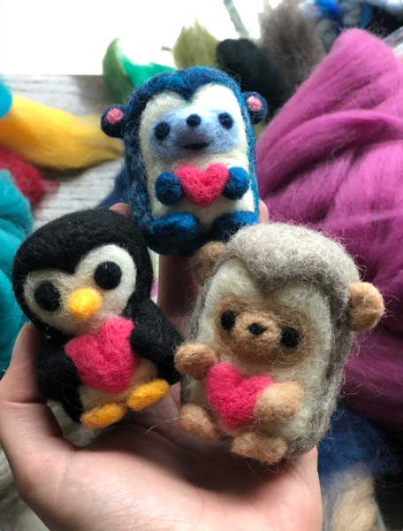 closeup of 3 felted animals in a hand: a penguin a blue bear and a hedgehog all holding felted hearts