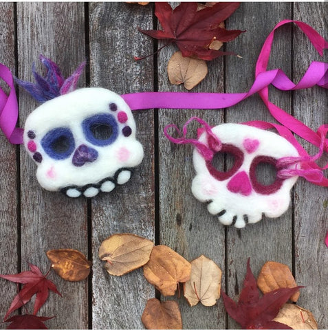 2 sugar skulls masks with ribbons to tie the ends. The left mask is white with purple and pink accents and the mask on the right is point. The masks are on a wooden deck with autumn leaves surrounding them and the photo is shot from overhead.