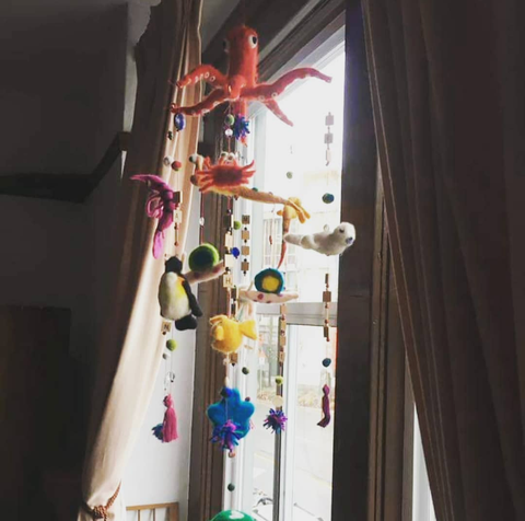 a felted baby mobile hanging in a window. A red octopus with strings hanging from each of its tentacles. On each string hangs a string with other sea creates and scrabble letters