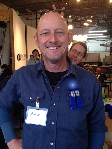 a male light skinned teacher with a shaved head is wearing a blue work jumpsuit. He is smiling with a name badge that says Roger on the left side of his chest and a light up badge of a blue tardis from Dr Who