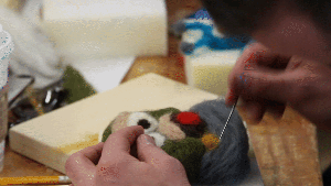 the needle felting process: closeup of a hand poking wool with a barbed needle repeatedly. The wool is shaped like a green monster cartoon character