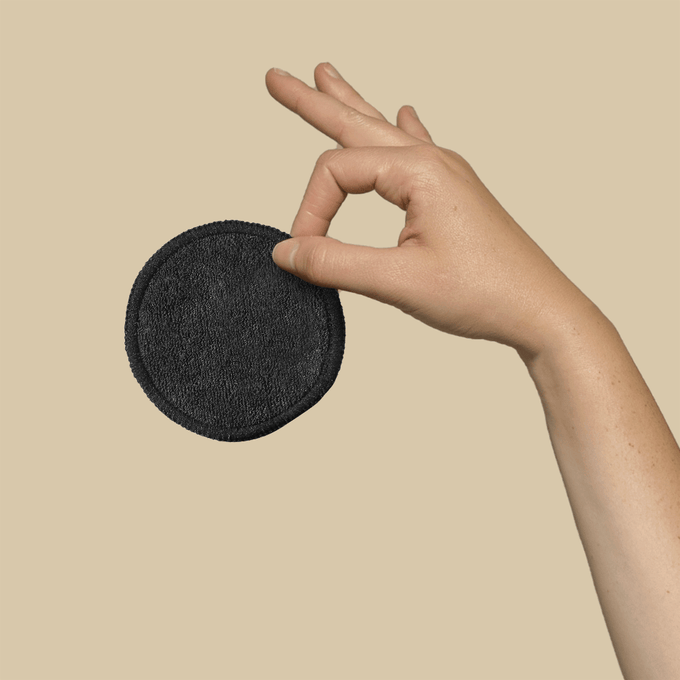A hand holds a single black bamboo cotton rounds against a neutral background. 