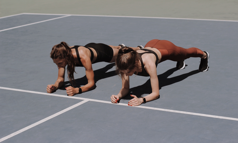 two women doing an elbow plank side by side on a track field