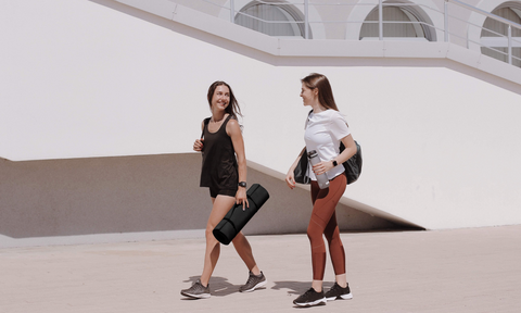 two women laughing and walking side by side, one is holding a yoga mat while the other is holding a water bottle