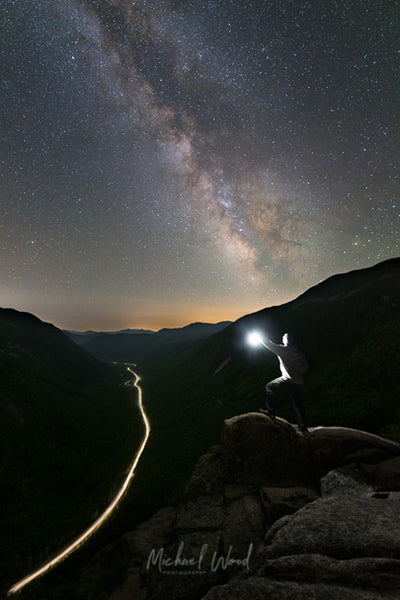 A hiker stands on top of a boulder as the Milky Way core rises over Crawford Notch in the White Mountains of New Hampshire