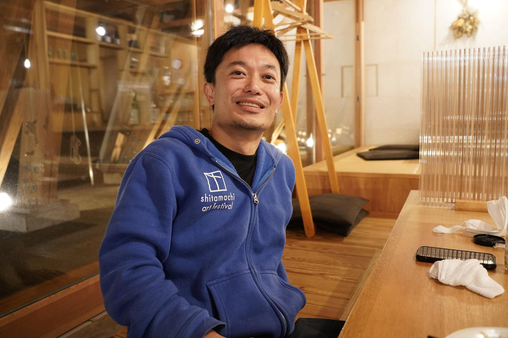 笑顔でカメラを見る渡辺さん、青いパーカーをきて、カフェのようなところに座る
