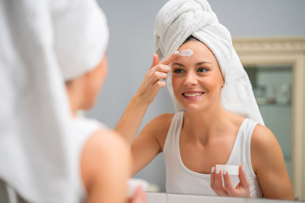 middle aged woman applying cream to her face while wearing towel on her hair