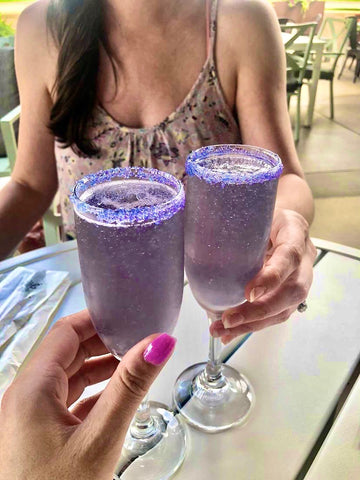 Two ladies holding champagne flutes with Lavendar French 75 cocktails embellished with Sugar Mama Shimmer edible drink glitter in Little Lilac and Violet Vibes. The champagne flute is rimmed in sugar and Sugar Mama Shimmer edible glitter for drinks.